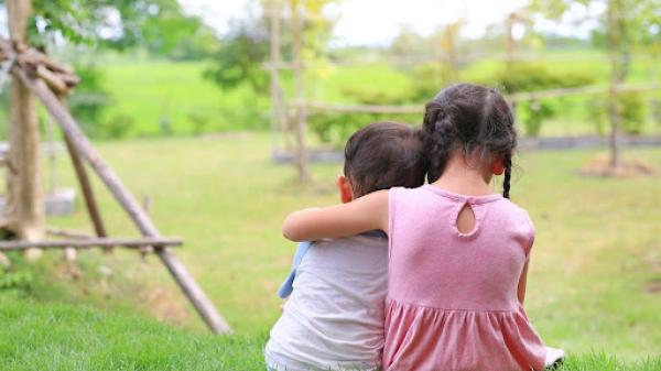 older child with arm around younger child on a green lawn